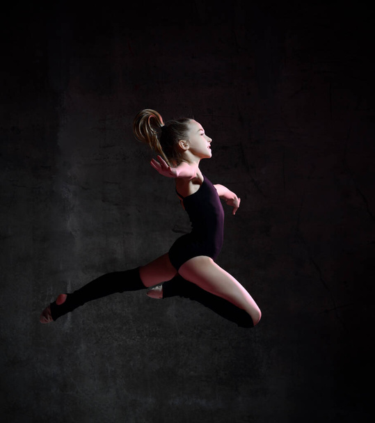 Young girl gymnast in black sport body and special uppers jumping in gymnastic pose over dark background - Foto, Bild