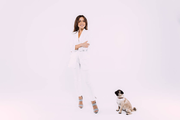 A laughing and smiling auburn haired woman in a white dress and white shoes, is staring most lovingly at her cute pug, who calmly sits down, gaining her undivided attention. Isolated white background - Photo, image