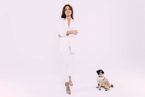 A laughing and smiling auburn haired woman in a white dress and white shoes, is staring most lovingly at her cute pug, who calmly sits down, gaining her undivided attention. Isolated white background - Foto, Bild