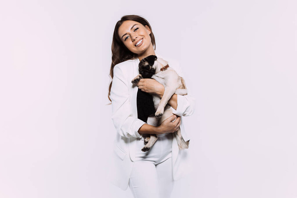 A laughing and smiling auburn haired woman in a white dress, is staring most lovingly at her cute pug, who calmly sits on the hands, gaining her undivided attention. Isolated white background - Photo, Image