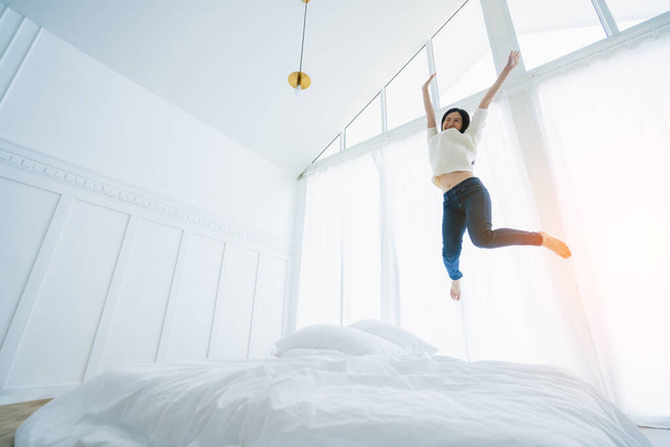 exited asian woman jump over white bed in bedroom with energy and happiness success lifestyle ideas concept - Foto, Imagen
