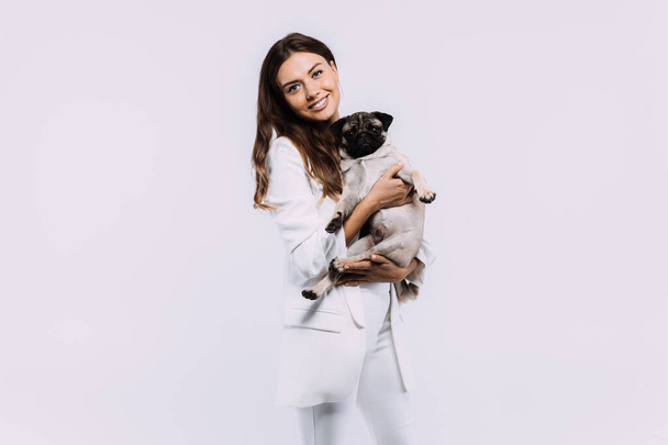 Μια γελαστή και χαμογελαστή μελαχρινή γυναίκα με λευκό φόρεμα, κοιτάζει με αγάπη το χαριτωμένο της σκυλάκι, που κάθεται ήρεμα στα χέρια, κερδίζοντας την αμέριστη προσοχή της. Μεμονωμένο λευκό φόντο - Φωτογραφία, εικόνα