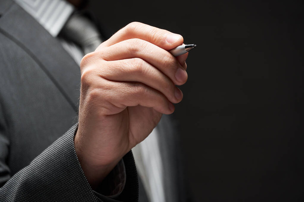 a pen closeup in businessman hand, gray suit, dark wall background - Фото, изображение