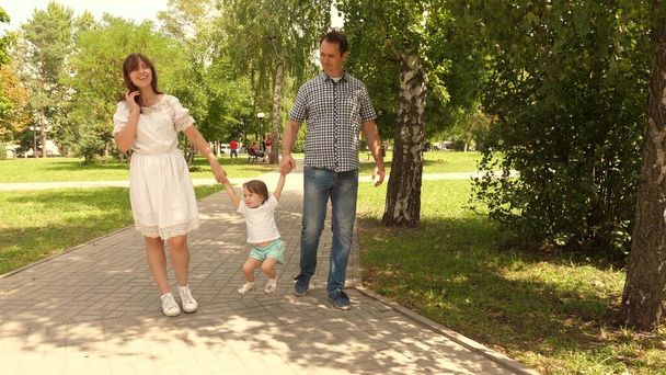 幸せな子供は父と母と遊ぶ手とジャンプを保持します。娘は公園でパパとママの手を握って飛び跳ねてる。家族の概念。自然の中で小さな子供と歩く. - 写真・画像