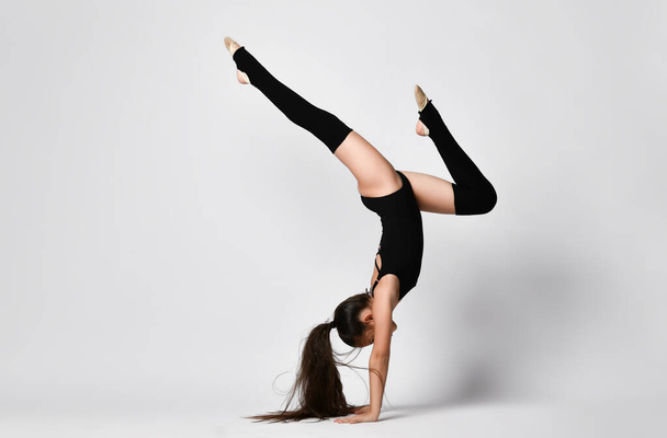 Teenage gymnast in black leotard, knee socks and ballet shoes, performing exercises standing on her hands, upside down, isolated on white. Close-up - Zdjęcie, obraz