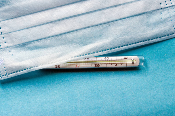 Μια μπλε ιατρική μάσκα προσώπου μιας χρήσης για την προστασία των ανθρώπων από έναν επικίνδυνο ιό, βρίσκεται στο παρασκήνιο της χρωματικής τάσης 2020. Υγειονομική περίθαλψη και ιατρική αντίληψη για το κινέζικο ξέσπασμα του coronavirus. θερμόμετρο δείχνει υψηλή θερμοκρασία - Φωτογραφία, εικόνα