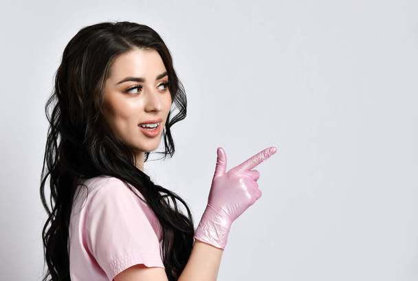 Girl in pink uniform and disposable gloves standing sideways, isolated on white. She smiling, pointing at something. Close up - Фото, зображення