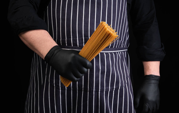 σεφ σε μαύρα γάντια λάτεξ, ριγέ ποδιά κρατά ακατέργαστο μακρύ κίτρινο σπαγγέτι από σκληρό σιτάρι, κοντά - Φωτογραφία, εικόνα