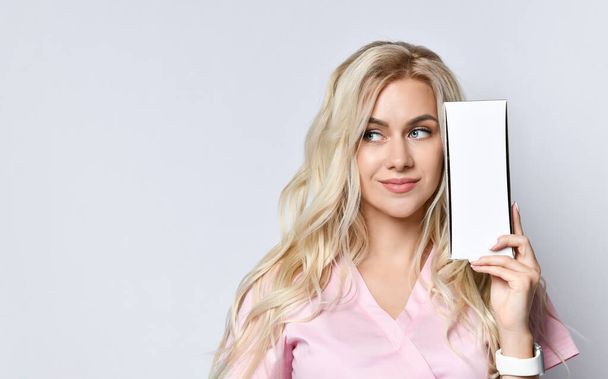 Blond woman with lovely makeup posing isolated on white. Smiling, holding a package of cosmetic product near the face with copy space for your text, logo. - Фото, зображення