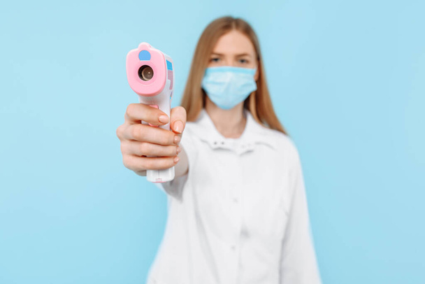 a female doctor, wearing a medical mask and white coat, uses a non- contact infrared forehead thermometer to check the patient's body temperature for symptoms of the virus against a blue background. - Zdjęcie, obraz