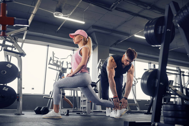 Side view of focused and motivated sporty young girl in sportswear doing legs exercises while handsome muscular personal trainer monitoring her . - Foto, afbeelding