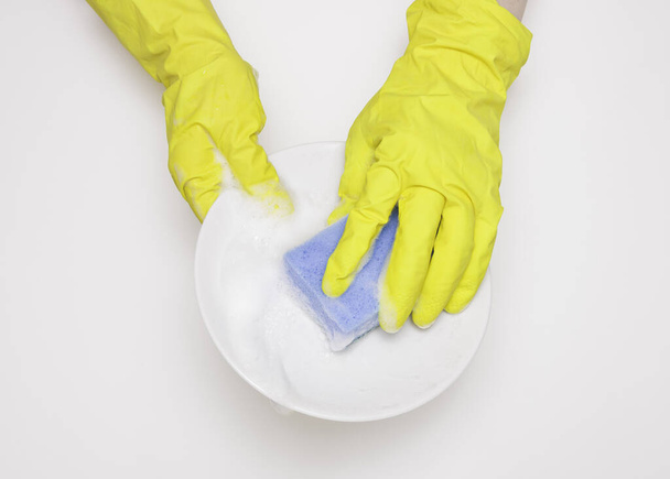 Hände in Gummihandschuhen verschließen den Teller mit Schwamm und Schaumstoff. Spülmittel, sauber. Weißer Hintergrund - Foto, Bild