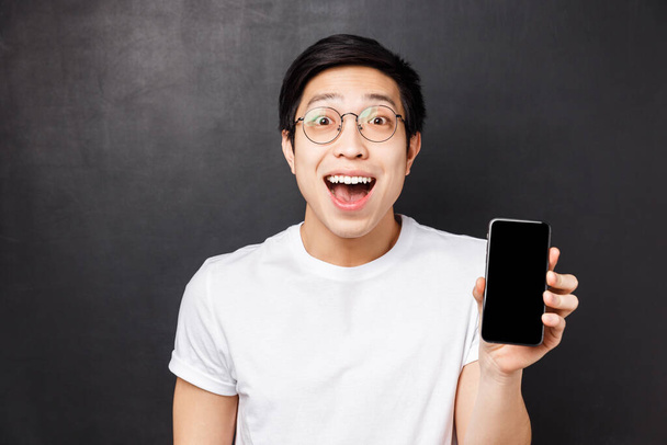 Τεχνολογία, μηνύματα και οι άνθρωποι έννοια. Κοντινό πλάνο πορτρέτο του χαρούμενος, έκπληκτος και εντυπωσιασμένος νεαρός ενθουσιασμένος Ασιάτης άνδρας σε λευκό t-shirt, δείχνει οθόνη smartphone έκπληκτος με δροσερό app - Φωτογραφία, εικόνα