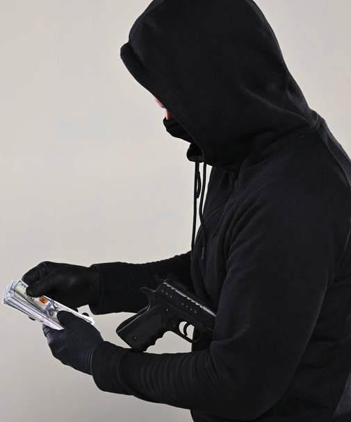 Man in black mask and hoody with a gun and pack of dollars over gray background  - Foto, afbeelding