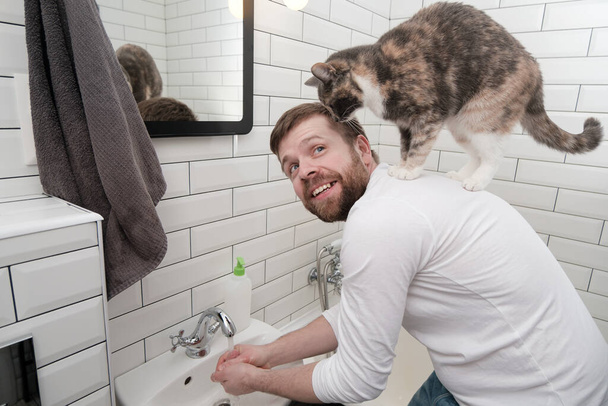 Satisfied man washes his hands, under a stream of water and his adorable cat climbed onto his back in the bathroom. - Fotó, kép