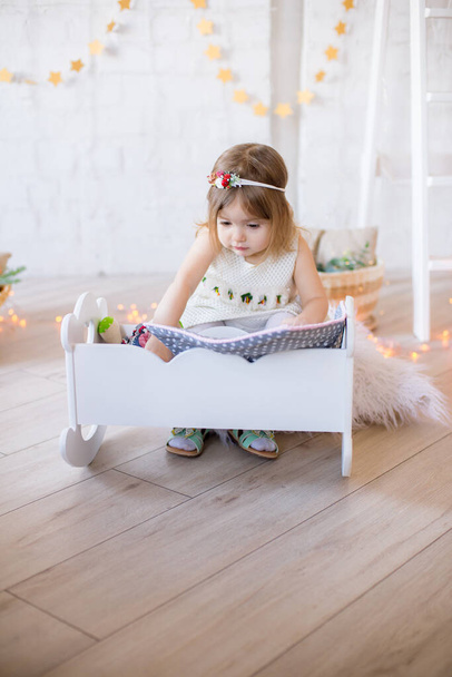 Little cute girl in a white dress plays in a doll bed in a bright children's room, decorated with toys and garlands. - Foto, immagini