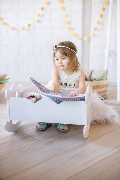 Little cute girl in a white dress plays in a doll bed in a bright children's room, decorated with toys and garlands. - Photo, image