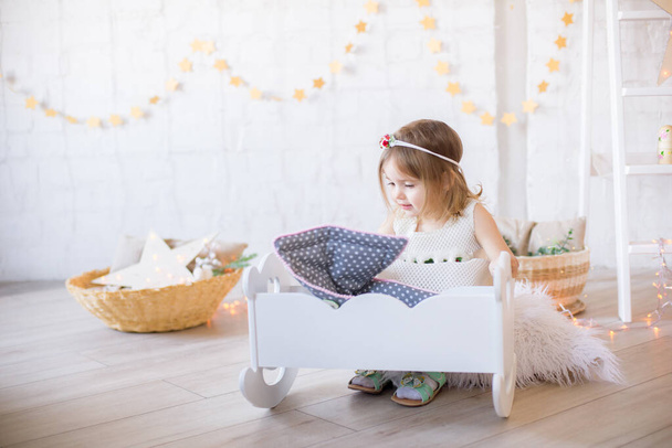 Little cute girl in a white dress plays in a doll bed in a bright children's room, decorated with toys and garlands. - Foto, Imagem