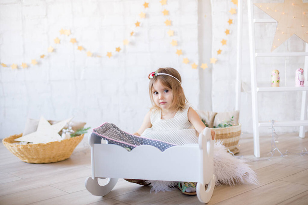 Little cute girl in a white dress plays in a doll bed in a bright children's room, decorated with toys and garlands. - Foto, imagen