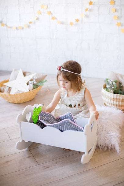 Petite fille mignonne dans une robe blanche joue dans un lit de poupée dans une chambre d'enfants lumineuse, décorée avec des jouets et des guirlandes
. - Photo, image