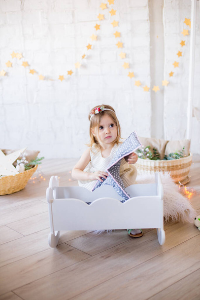 Little cute girl in a white dress plays in a doll bed in a bright children's room, decorated with toys and garlands. - Photo, image