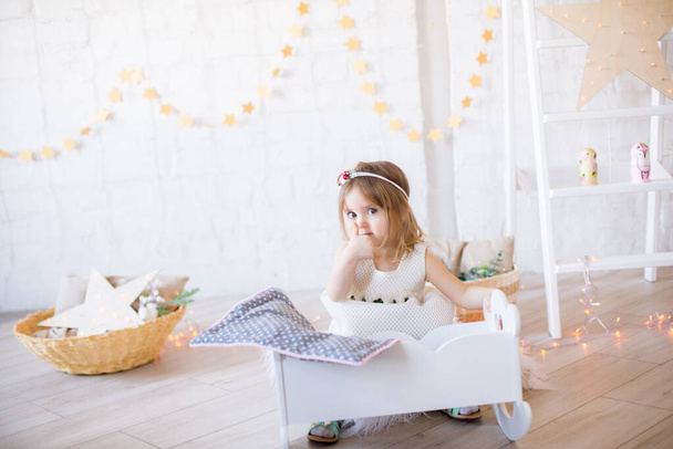 Little cute girl in a white dress plays in a doll bed in a bright children's room, decorated with toys and garlands. - Foto, Bild