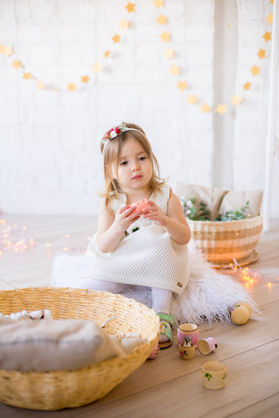 Little cute girl in a white dress plays in a bright children's room, decorated with toys and garlands. - Fotó, kép