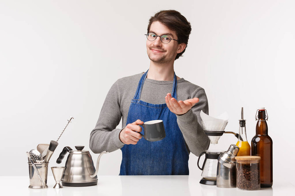 Barista, cafe worker and bartender concept. Here you are. Charismatic friendly young bearded male employee made coffee for customer, giving cup of cappuccino, standing white background - Valokuva, kuva