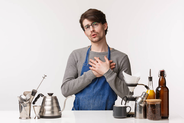 Barista, cafe worker and bartender concept. Portrait of touched handsome young male employee in apron making coffee, touch heart and feel flattered, customer likes his cappuccino - Photo, image