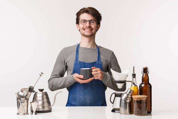 Barista, cafe worker and bartender concept. Portrait of cheerful pleasant young male employee in apron invite everyone have delicious drink, making coffee, holding tea and smiling camera - Photo, Image