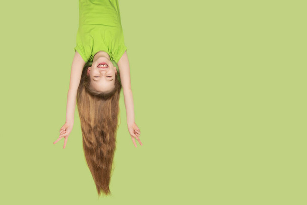 Grappig en mooi kind meisje met extreem lang haar glimlachend geïsoleerd op groene achtergrond. Meisje dat ondersteboven hangt met vliegend haar. Kopieerruimte. - Foto, afbeelding