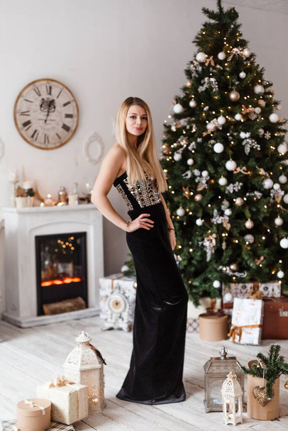A luxurious blonde in a black velvet evening long dress stands at the decorated Christmas tree and smiling, preparing to celebrate the New Year and Christmas - Фото, изображение
