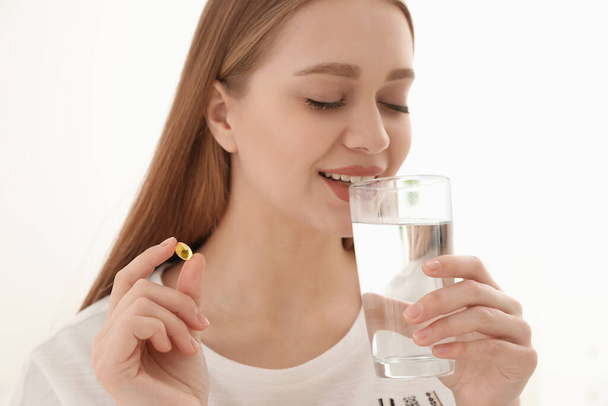 Young woman with glass of water taking vitamin capsule on light background - Foto, Imagen