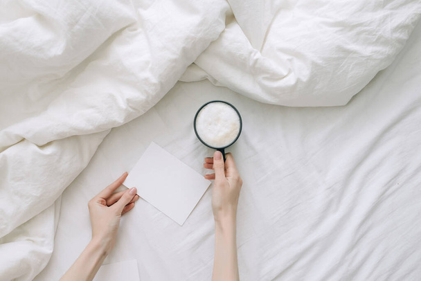 Female hand holding coffee cup and postcard in white bedclothes. Top view.  - Photo, Image