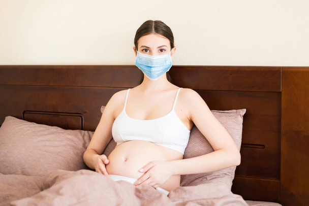 Koruyucu tıbbi maskeli hamile kadın koronavirüs hastası ve yataktan çıkmıyor. Fetüs enfeksiyonunu önlemek için. Küresel salgın. Karantina konseptini koruyun.. - Fotoğraf, Görsel
