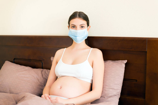 Nuori raskaana oleva nainen suojaava lääketieteellinen naamio pysyy sängyssä, koska sepelvaltimovirus. Karanteeniaika. Terveydenhuollon käsite
. - Valokuva, kuva