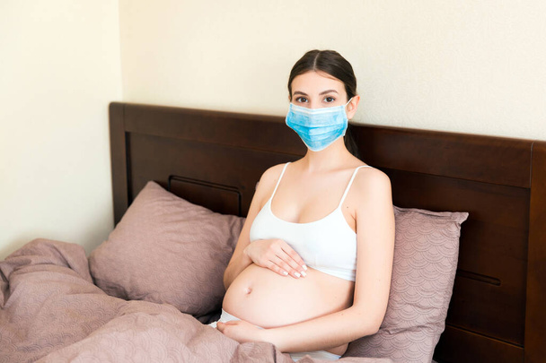 Young pregnant woman in protective medical mask is staying in bed because of coronavirus. Quarantine time. Health care concept. - Foto, imagen