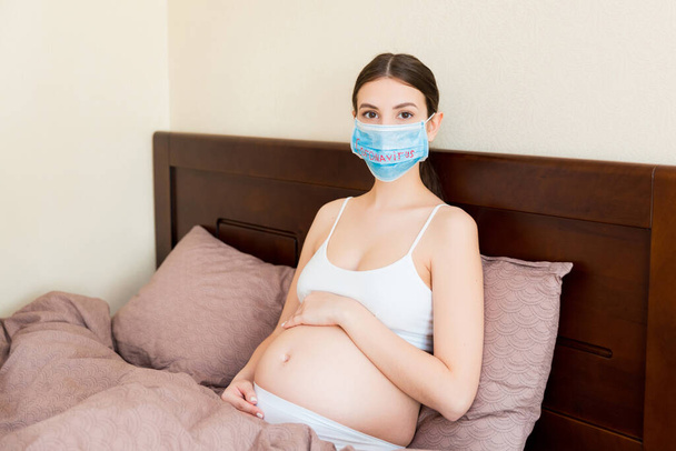 Έγκυος γυναίκα με προστατευτική ιατρική μάσκα με coronavirus λέξη μένει στο κρεβάτι. Επιδημικό ξέσπασμα. Παγκόσμια καραντίνα. - Φωτογραφία, εικόνα