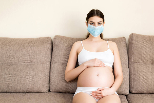 妊娠中の女性は自分自身と将来の子供を医療マスクでコロナウイルス病から守っています。お母さんは家でソファーに座っている。流行概念. - 写真・画像