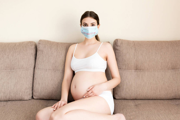 Kranke Schwangere sitzt zu Hause auf der Couch. Schützen Sie Ihre Gesundheit mit einer medizinischen Schutzmaske. Coronavirus-Infektion. Globale Quarantäne. - Foto, Bild