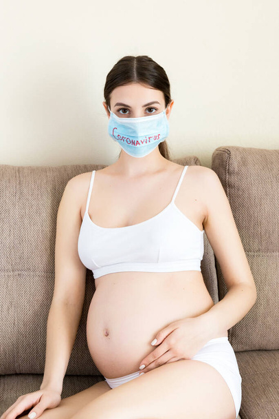 妊婦は自宅のソファに座っている。保護医療マスクであなたの健康を守る。コロナウイルス感染症。全球隔離. - 写真・画像