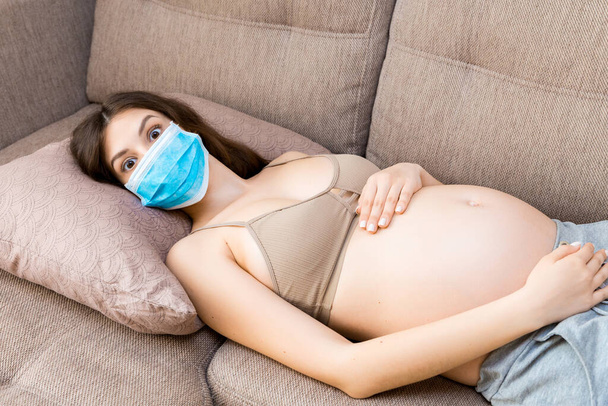 Έγκυος γυναίκα προστατεύει τον εαυτό της και το μελλοντικό παιδί από την ασθένεια coronavirus με ιατρική μάσκα. Σοκαρισμένη μαμά είναι ξαπλωμένη στον καναπέ στο σπίτι. Έννοια επιδημικής έξαρσης. - Φωτογραφία, εικόνα