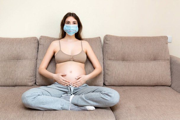 Kobieta w ciąży w ochronnej masce medycznej ze słowem koronawirus siedzi na kanapie w domu z powodu choroby układu oddechowego. Zatrzymanie globalnej koncepcji epidemii. - Zdjęcie, obraz