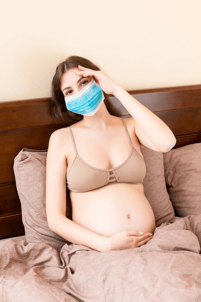 保護医療マスクの若い妊婦は頭痛に苦しんでおり、コロナウイルスのためにベッドに滞在しています。隔離時間だ。ヘルスケアの概念. - 写真・画像