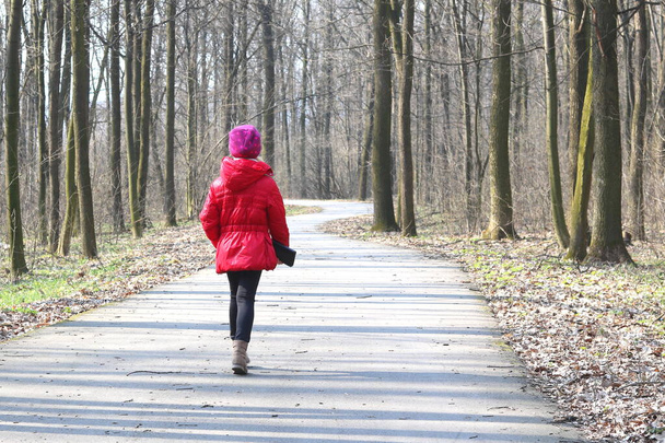 Little girl in red jacket and red hat walks in spring forest among trees on asphalt road - Foto, Imagen