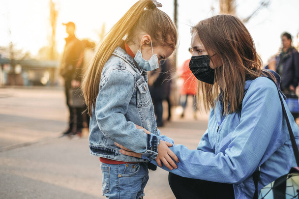 Ένα κορίτσι με ένα παιδί στέκεται στο δρόμο με μια προστατευτική ιατρική μάσκα. Επικίνδυνη λοίμωξη από ιό. Ο κορονοϊός στην Ευρώπη - Φωτογραφία, εικόνα