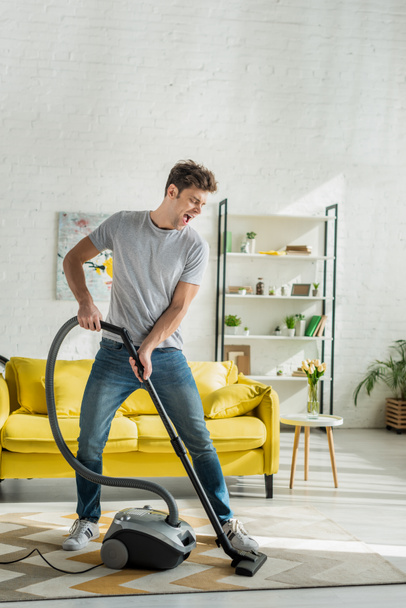 handsome man with opened mouth using vacuum cleaner in living room  - Fotoğraf, Görsel
