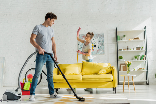 happy man using vacuum cleaner near woman waving hand in living room  - Foto, imagen