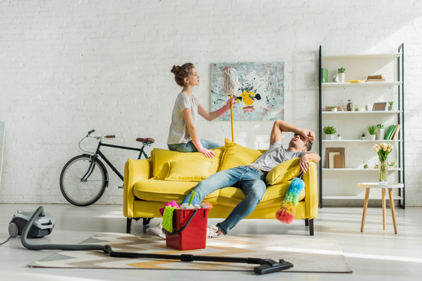 exhausted man sitting on sofa near bucket with household and woman  - Photo, Image