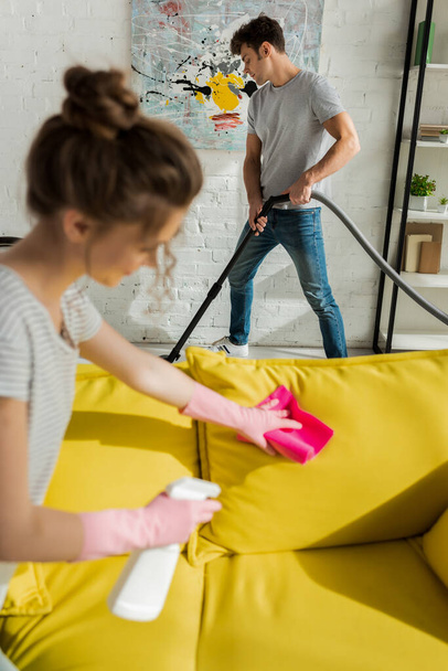 selective focus of handsome man using vacuum cleaner near girl washing sofa with rag  - Фото, изображение